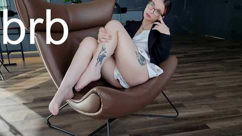 Media: A video of a fair-skinned woman with glasses, tattoos, and short hair, wearing a white dress and black jacket, lounging on a brown leather chair in a modern room with wooden floors.