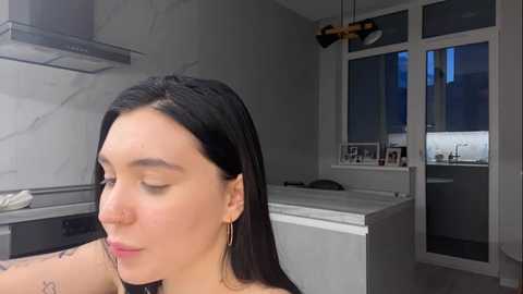 Media: A video of a fair-skinned woman with long black hair, wearing minimal makeup, in a modern, minimalist kitchen. She has a nose ring and tattoos on her arm.