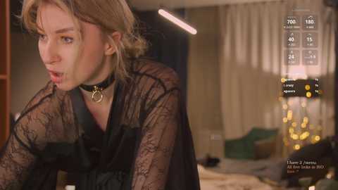 Media: Video of a blonde woman with a shocked expression, wearing a black lace dress and choker, in a dimly lit bedroom with a bed and curtains in the background.