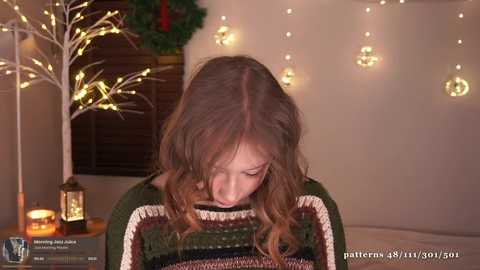 Media: Video of a young woman with long, wavy brown hair wearing a green, hand-knit sweater, looking down in a cozy room with Christmas decorations, including a tree, lights, and lanterns.