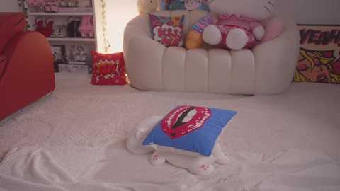 Media: Video of a plush toy dog wearing a blue pillow with a red lipstick print, lying on a white carpeted floor in a pink-themed room with cartoon posters and plush toys on shelves.