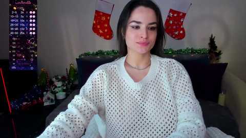 Media: Video of a young woman with long dark hair, wearing a white knitted sweater, sitting on a bed with Christmas decorations in the background.