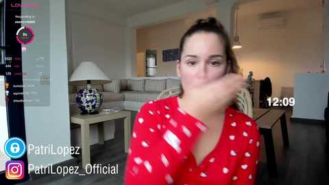 Video of a young woman with brown hair in a ponytail, wearing a red blouse with white heart patterns, sitting in a modern living room with a couch, lamp, and a TV screen displaying \"12:09\" and \"PartiLopez Official.\