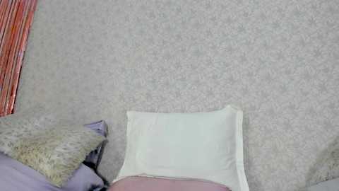 Media: Video of a cozy bedroom with a beige carpeted floor and a white pillow on a bed. A soft, light purple blanket is draped over the pillow, and a pink shirt is visible in the lower part of the image.