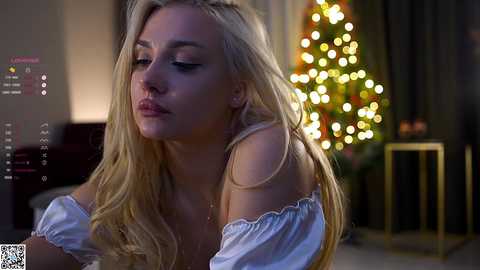 Media: A video of a blonde woman with long hair, wearing a white off-shoulder top, gazing thoughtfully at a Christmas tree in a dimly lit room.