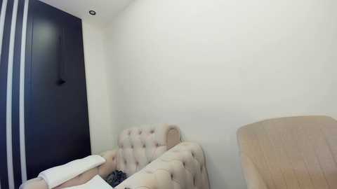 Media: Video of a minimalist, modern living room corner with beige tufted sofa, white wall, and wooden chair, featuring a flat-screen TV and a decorative black wall hanging.