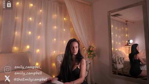 Media: Video of a woman with long black hair, wearing a black dress, seated at a table in a softly lit room with string lights and a mirror.