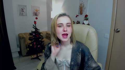 Media: Video of a blonde woman in a patterned robe, smiling and sticking out her tongue, in a cozy living room with a Christmas tree, beige couch, and festive decorations.