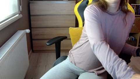 Media: A video of a woman with medium-length brown hair, wearing a light pink sweater and grey pants, sitting in a yellow gaming chair in a wooden-furnished room.