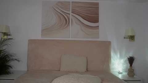 Media: Video of a minimalist bedroom with a beige headboard, a textured cream rug, abstract art above, white wall-mounted sconces, and a potted plant on the left.