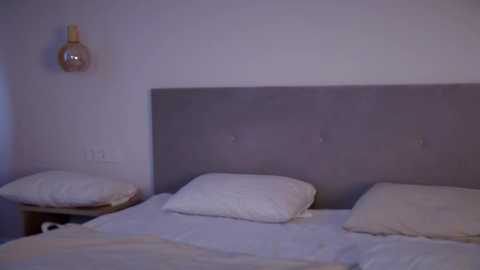 Media: Video of a minimalist bedroom with a gray upholstered headboard, two white pillows, and a nightstand with a lamp. The walls are painted white, creating a serene and modern atmosphere.