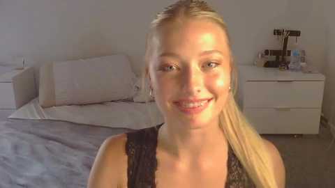 Media: Video of a smiling young blonde woman with straight hair tied back, wearing a black lace top, standing in a modern bedroom with a white bed, dresser, and camera equipment.