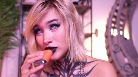 Media: Video of a blonde woman with blue eyes and black lipstick, eating a carrot, wearing dark makeup, and a black top. She has intricate tattoos on her neck and chest. Background includes a plant and ornate mirror.