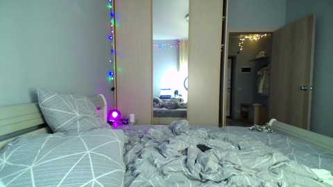 Media: Video of a messy, cozy bedroom with a bed covered in light gray bedding, a tall mirror with colorful string lights, and an open door revealing a small, cluttered bathroom.