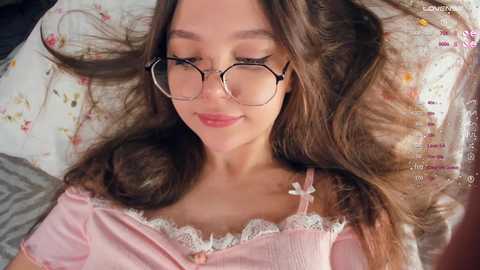 Media: A young woman with fair skin and long brown hair lies on a floral-patterned pillow, wearing glasses and a pink, lace-trimmed top.