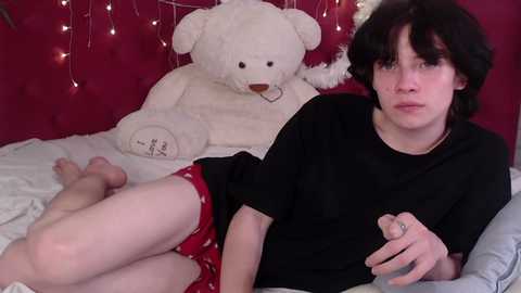 Video of a young person with short black hair, wearing a black shirt and red shorts, lying on a bed with a white bear and red string lights in the background.