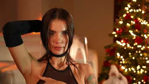 Media: Video of a woman with long brown hair, wearing a black halter top and black elbow-length gloves, posing in front of a decorated Christmas tree.