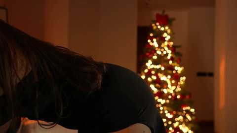 A video captures a person's back, wearing a black shirt, in a warmly-lit room with a Christmas tree adorned with red and gold ornaments and twinkling lights.