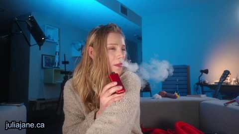 Media: Video of a blonde woman in a beige sweater, exhaling smoke from a red cigarette, indoors under blue lighting, with a studio backdrop, by julienne.ca.