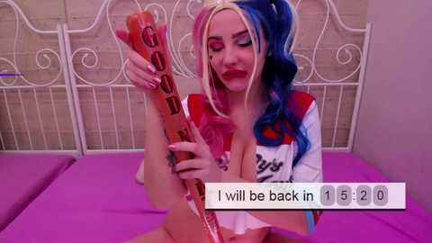Video of a woman with pink and blue hair, wearing a Harley Quinn top, holding a bat, lying on a bed with purple sheets, in a bedroom.