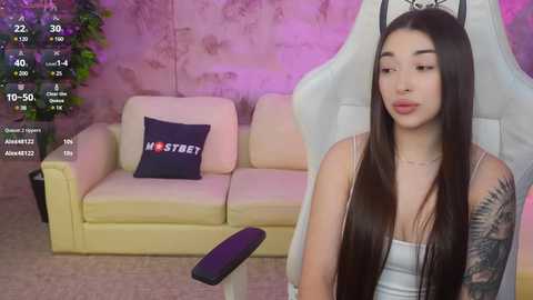 Media: Video of a young woman with long dark hair, wearing a white tank top, seated on a white gaming chair in a room with a beige couch and a \"ME STREET\" pillow.