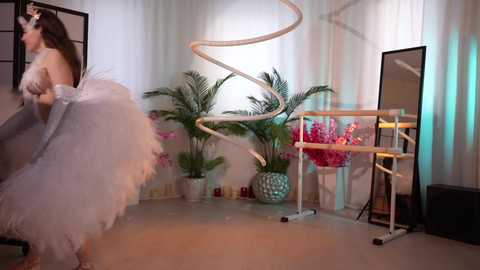 Media: A video of a woman in a voluminous white dress, with a curved mirror, greenery, and pink flowers in a modern, minimalist room.