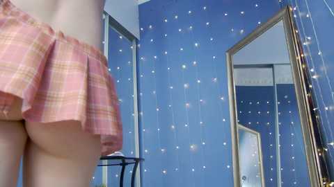 Media: Video of a fair-skinned woman wearing a pink plaid mini-skirt, standing in a room with blue walls adorned with fairy lights. The image focuses on her lower back and skirt, capturing a casual, intimate moment.