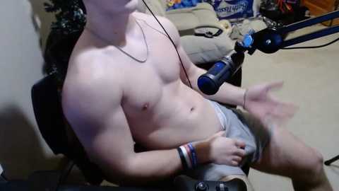 Media: Video of a shirtless, muscular man with fair skin, sitting on a black chair, wearing gray shorts, recording himself with a blue microphone on a stand. Background includes a beige couch, carpeted floor, and a blue wall.