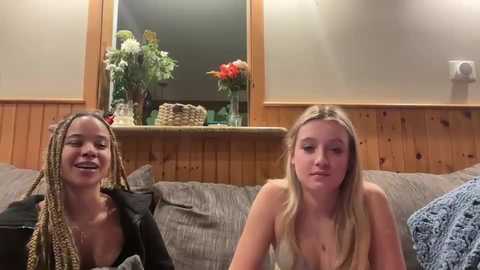 Media: Video of two young women, one with braided hair, the other with long straight hair, seated on a couch, smiling, in a cozy, wooden-paneled room with a floral arrangement on a ledge above them.