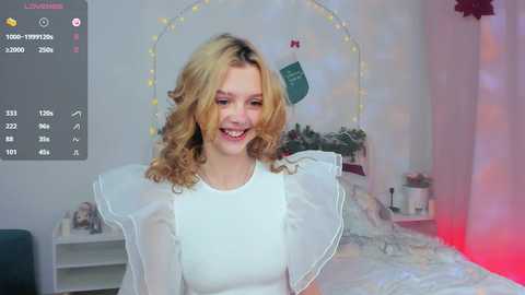 Media: Video of a smiling young woman with wavy blonde hair, wearing a white blouse, standing in a softly lit, cozy bedroom with fairy lights and plants.