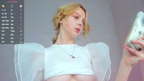 Media: A video of a fair-skinned, blonde woman with wavy hair, wearing a white crop top with sheer sleeves, looking at a smartphone. Background is a plain, light-colored wall.