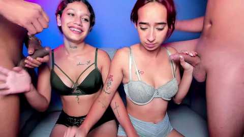 Media: Video of two young women with short hair, tattoos, and light brown skin, wearing lingerie, surrounded by multiple erect penises.