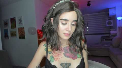 Media: Video of a young woman with long wavy dark hair, wearing a black lace bralette, showcasing colorful floral tattoos on her chest, standing in a dimly lit room with posters on the wall and a couch.