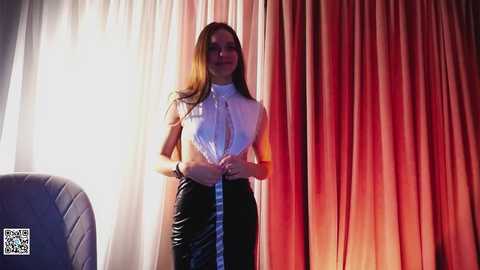 Media: Video of a slim, fair-skinned woman with long brown hair, wearing a white blouse and black skirt, standing against a gradient curtain backdrop in a modern indoor setting.