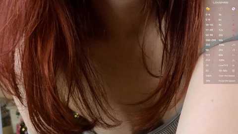 Close-up video of a woman with medium-length, straight, reddish-brown hair, wearing a low-cut, grey shirt, revealing cleavage. Background includes a digital calendar with daily tasks and appointments.