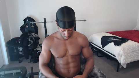 Media: Video of a shirtless, muscular Black man with a black baseball cap, standing in a sparsely furnished room with a white bed, red blanket, and black bags.