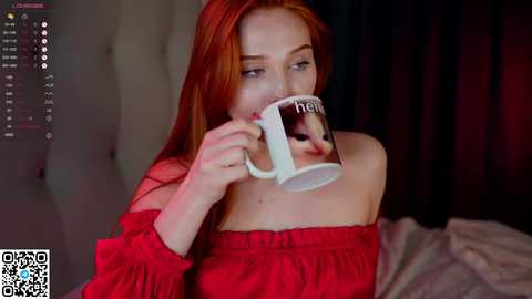 Media: Video of a fair-skinned woman with long, straight red hair, wearing a red off-shoulder dress, drinking from a \"hello\" mug, in a dimly lit room with a bed in the background.