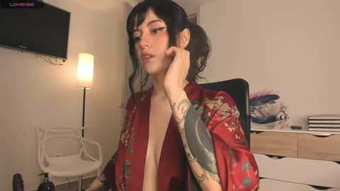 Media: A young woman with pale skin, dark hair, and tattoos sits in a dimly lit room, wearing a red kimono and a contemplative expression.