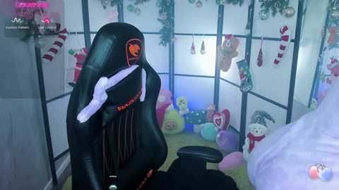 Media: Video of a gamer in a black gaming chair with orange accents, surrounded by festive decorations including Christmas trees, presents, and colorful balloons in a room with white walls and black curtains.