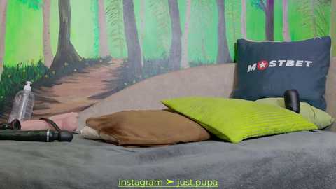 Media: Video of a gray bed with a green pillow and a black pillow with \"MOSTBET\" logo. A bottle of lubricant, a dildo, and a condom are on the bed. Background features a green forest mural.