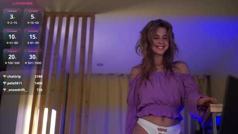 Media: A video of a young woman with long, wavy brown hair, smiling in a purple off-shoulder top, white panties, standing in a dimly lit room with wooden blinds and blue lighting.