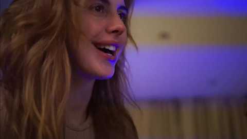 Media: Video of a young Caucasian woman with long, wavy blonde hair, smiling, with a hint of blue light in the background, wearing a brown jacket.