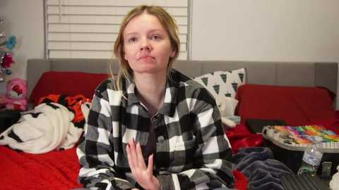 Media: Video of a blonde woman in a black-and-white checkered shirt, sitting on a messy bed with red sheets, surrounded by scattered clothes and a colorful candy bowl.