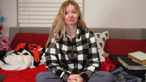 Media: Video of a smiling, light-skinned blonde woman in a black-and-white checkered shirt and gray pants, sitting on a red bed with a pile of clothes, a book, and a stuffed animal.