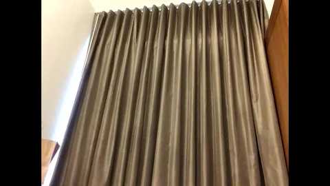 Media: Video of a room with a large, heavy, gray satin curtain covering the window, creating a luxurious and sophisticated ambiance. The curtain's texture is smooth and reflective. The room has white walls and a wooden chair on the left side.