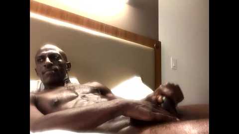 Media: Video of a shirtless Black man with a shaved head, lying on a bed, holding a large, erect penis. The background shows a wooden headboard and beige walls.