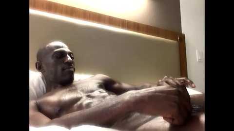 Media: A video of a muscular, bald Black man lying on a bed, shirtless, with a relaxed expression. The room has a wooden headboard and beige walls.