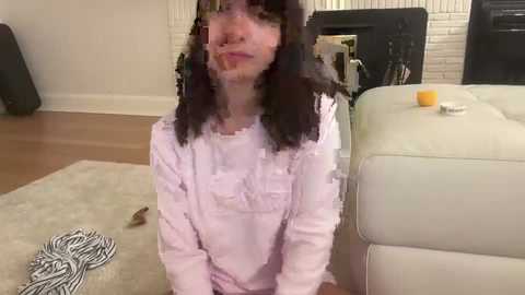 Media: A video of a young woman with curly black hair, wearing a white shirt, kneeling on a beige carpet, next to a white couch, in a modern living room.