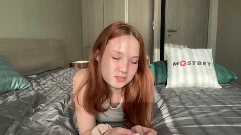 Media: Video of a young woman with fair skin and long, straight red hair lying on a bed in a bedroom, wearing a white tank top, with a \"MOSTBET\" pillow in the background.