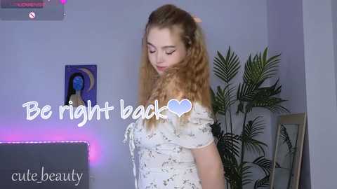 Media: Video of a young woman with long, curly blonde hair, wearing a white floral dress, standing in a room with purple lighting, a potted plant, and framed artwork. Text reads \"Be right back \u2665 cute beauty.\